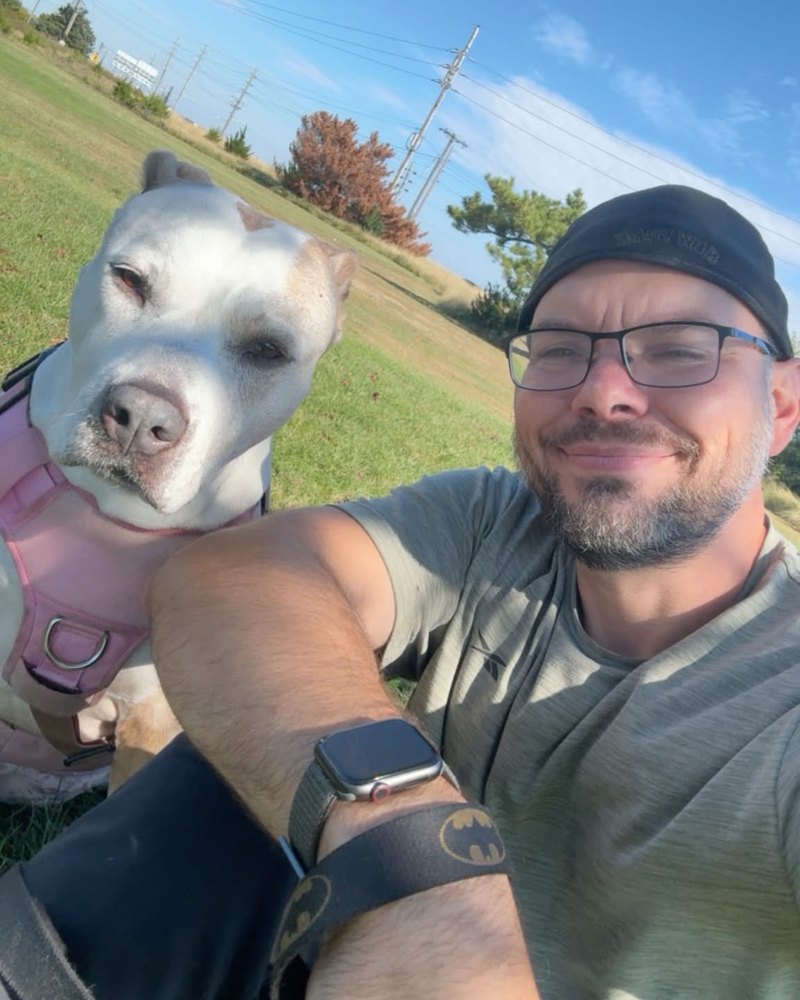 James smiling in a cap and glasses, sitting in the grass on a sunny day next to Seven, a white dog in a pink harness