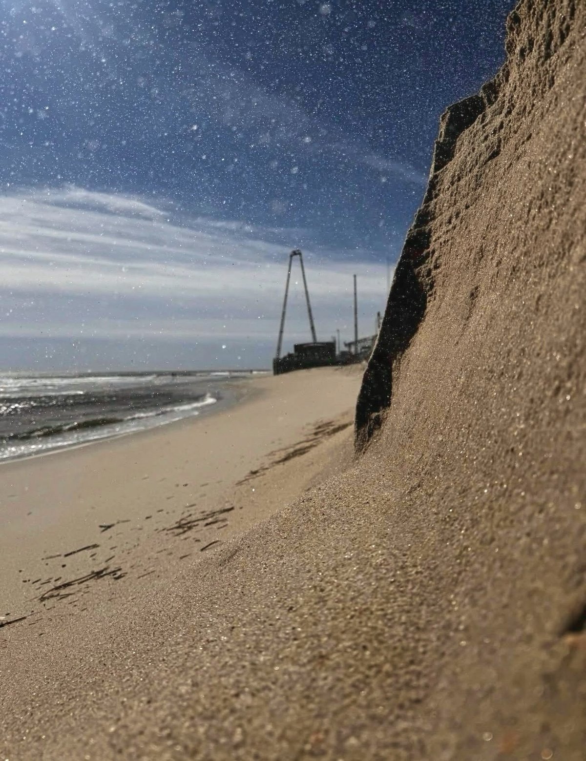 Low angle view along the beach at Seaside Heights, New Jersey: a sand bank rising on the right, surf on the left, an amusement pier ride in the distance and a deep blue sky streaked with cloud